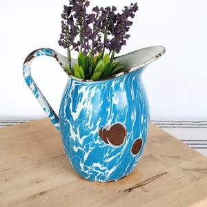 May include: A vintage blue and white enamelware pitcher with a marbled design. The pitcher has a handle and is filled with purple and green faux flowers. The pitcher is sitting on a wooden surface.