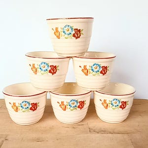 May include: A stack of six cream-colored ceramic bowls. Each bowl features a floral design with blue, red, and yellow flowers. The rims are trimmed with a thin red line. The bowls have horizontal ridges.