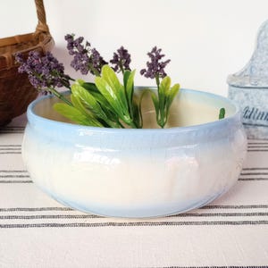 May include: A round, light blue and white ceramic bowl with a crackle glaze, filled with purple and green faux flowers. A woven basket and a blue wall-mounted container are in the background.