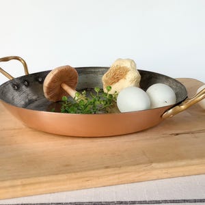 May include: A round copper pan with two brass handles, featuring a dark metallic interior. Inside the pan are a wooden mushroom figurine, a small bunch of green herbs, and two white eggs, with an additional white egg resting outside the pan.