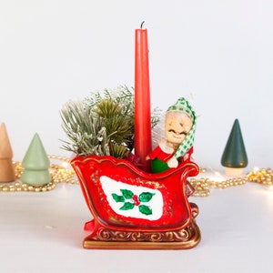 May include: A red ceramic sleigh candle holder featuring a hand-painted holly leaf design on the side. The sleigh contains a tall red taper candle, artificial pine greenery, and a small elf figurine wearing a green and white checkered hat.