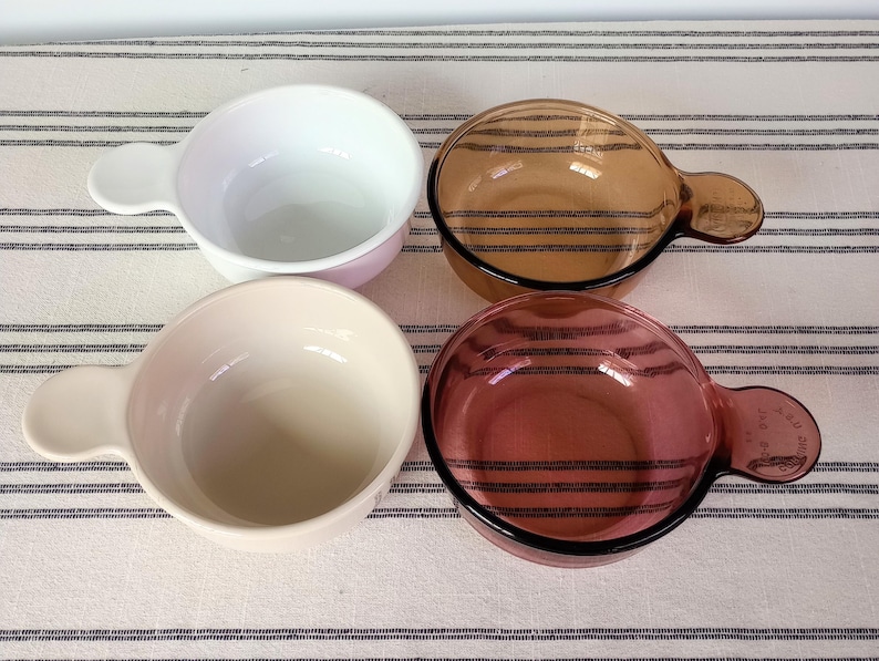 Pode incluir: Quatro tigelas de vidro com al&ccedil;as, em branco, rosa claro, &acirc;mbar e vermelho-p&uacute;rpura. As tigelas s&atilde;o redondas com uma al&ccedil;a de um lado. As tigelas &acirc;mbar e vermelho-p&uacute;rpura t&ecirc;m listras pretas. As tigelas est&atilde;o em uma superf&iacute;cie listrada branca e cinza.