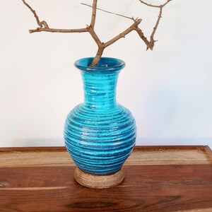 May include: A turquoise ceramic vase with a textured, ribbed design. The vase has a wide, round base and a flared top. A dried, brown twig is placed inside the vase. The vase is sitting on a wooden tray.