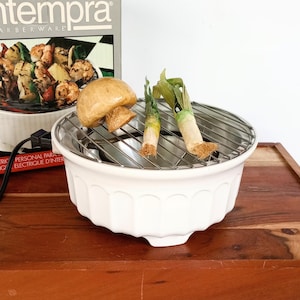 May include: A white ceramic electric grill with a circular metal grate, featuring a scalloped exterior design. The grill is positioned in front of its original box, which displays the brand name Contempra by Farberware and a food graphic.