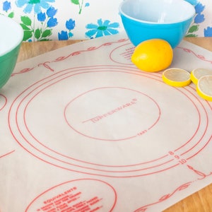 May include: A Tupperware baking mat with red circular guides and text, alongside a green and a blue bowl, and a whole lemon with lemon slices. The mat has measurements for tart sizes. The background features a floral wallpaper.