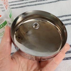 Puede incluir: Tapa de coctelera de acero inoxidable brillante, con un peque&ntilde;o orificio central. La tapa es redonda y reflectante. El texto "AIQUIRI" es visible en un vaso cercano.