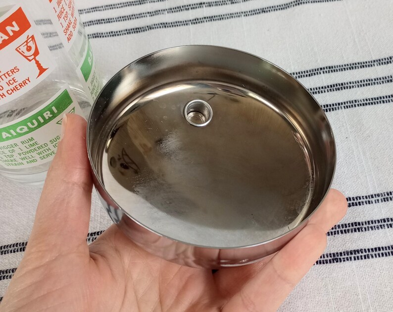 Puede incluir: Tapa de coctelera de acero inoxidable brillante, con un peque&ntilde;o orificio central. La tapa es redonda y reflectante. El texto "AIQUIRI" es visible en un vaso cercano.
