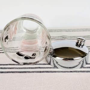 Puede incluir: Un vaso transparente con gr&aacute;ficos en blanco y negro y una petaca plateada con tap&oacute;n de rosca. El vaso est&aacute; tumbado de lado, revelando el dise&ntilde;o interior. La petaca es muy reflectante y el tap&oacute;n tiene textura.