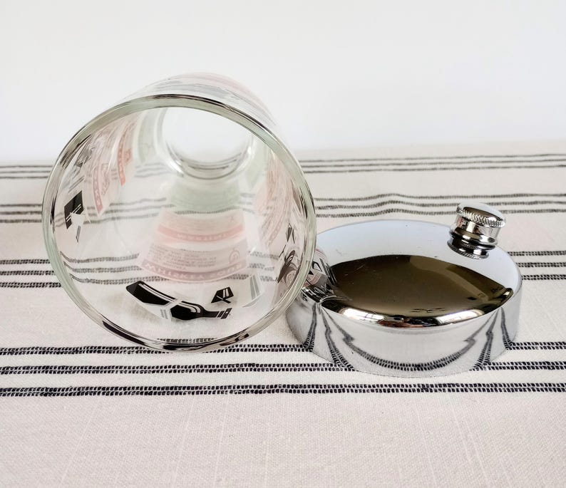 Puede incluir: Un vaso transparente con gr&aacute;ficos en blanco y negro y una petaca plateada con tap&oacute;n de rosca. El vaso est&aacute; tumbado de lado, revelando el dise&ntilde;o interior. La petaca es muy reflectante y el tap&oacute;n tiene textura.