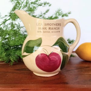 May include: Cream-colored ceramic pitcher with a handle and spout. The pitcher is decorated with a large red cherry and green leaves. Text on the pitcher reads "Lea Brothers Mink Ranch, Alma Center, Wisconsin."