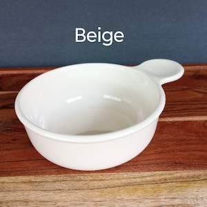 Pode incluir: Uma tigela de cer&acirc;mica bege com uma al&ccedil;a. A tigela &eacute; redonda com uma borda ligeiramente alargada e uma pequena al&ccedil;a arredondada que se estende da lateral. A palavra "Beige" &eacute; impressa em branco no canto superior esquerdo.