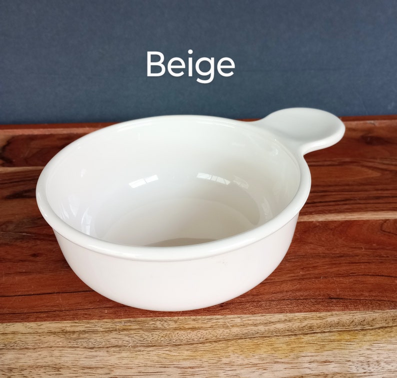 Pode incluir: Uma tigela de cer&acirc;mica bege com uma al&ccedil;a. A tigela &eacute; redonda com uma borda ligeiramente alargada e uma pequena al&ccedil;a arredondada que se estende da lateral. A palavra "Beige" &eacute; impressa em branco no canto superior esquerdo.