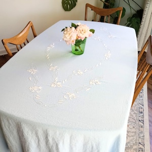 May include: A light blue oval tablecloth with embroidered floral detailing. A green glass vase holds a bouquet of pink roses. Wooden chairs surround the table, creating a vintage dining setting.
