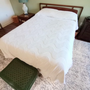 May include: A white bedspread with a wavy pattern covers a bed with a wooden headboard. A green ottoman with a patterned top sits at the foot of the bed. A fluffy white rug and a wooden nightstand are also visible.