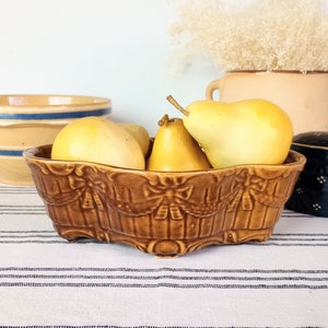 May include: A brown ceramic serving dish filled with yellow pears. The dish has a decorative embossed design. Other ceramic pieces are in the background, including a blue and tan bowl and a dark blue lidded container.
