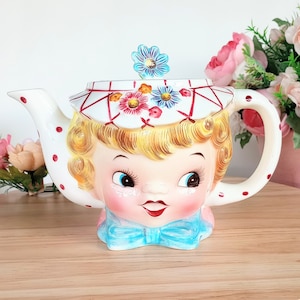 May include: A vintage-style ceramic teapot featuring a smiling girl's face. The teapot is white with red polka dots and a floral-decorated lid. The girl has blonde hair, blue eyes, and a blue bow tie. The handle is white with red polka dots.