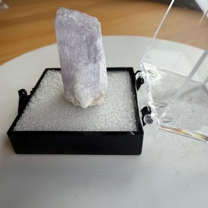 May include: A pale lavender, translucent mineral crystal standing upright on a white foam insert inside a small, square black display box with a clear, hinged plastic lid that is open to the right side, revealing the raw, textured stone surface.