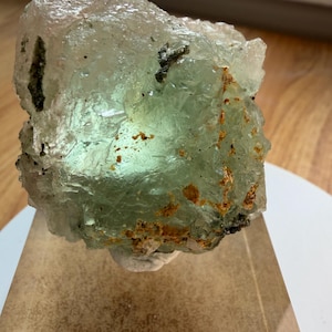 May include: A translucent, pale green fluorite crystal specimen with a rough, crystalline texture and visible internal fractures. The mineral features patches of orange-brown iron staining and dark inclusions, mounted on a clear acrylic block.