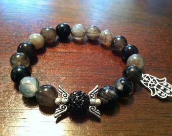 May include: A black and gray beaded bracelet with a silver charm featuring a hand with an eye in the palm. The bracelet has a silver charm with wings and a black crystal in the center.