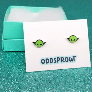 May include: A pair of green and black enamel earrings featuring a cartoon character with large ears. The earrings are displayed on a white card with the text "ODDSPROUT".