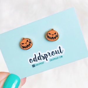 May include: A pair of gold-toned earrings featuring a jack-o'-lantern design. The earrings are on a white card with the text "oddsprout" and "oddsprout.com".
