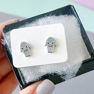 May include: A pair of silver stud earrings with a gray cupcake design. The cupcakes have a frowning face with two eyes and a mouth.