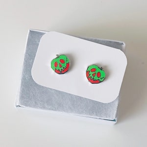 May include: A pair of silver earrings shaped like a green apple with a red bottom and a skull face. The earrings are in a white box.