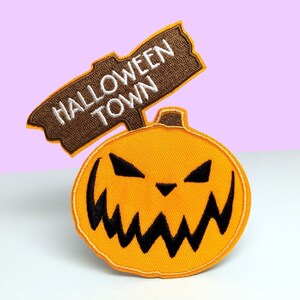 May include: An orange and black Halloween-themed iron-on patch featuring a jack-o'-lantern with a menacing grin and a brown sign that reads "Halloween Town".