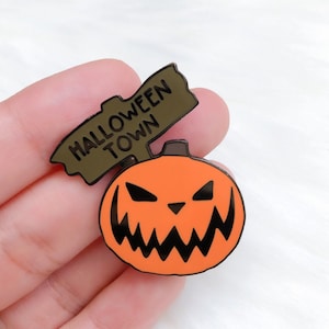 May include: A black and orange enamel pin featuring a jack-o'-lantern with a menacing grin and a wooden sign that reads "Halloween Town".