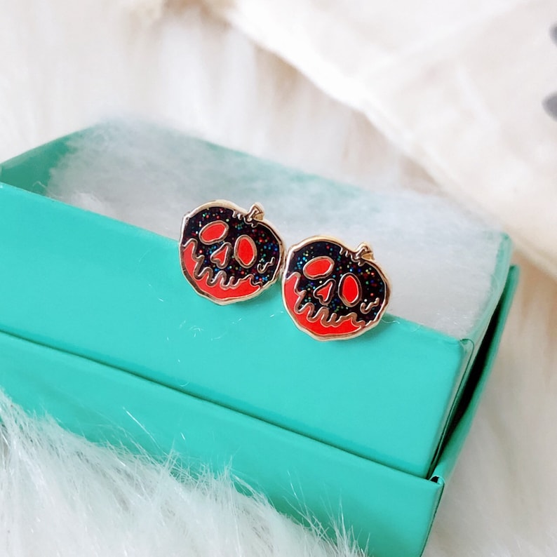 May include: A pair of gold-toned earrings featuring a red and black enamel design of a poisoned apple. The apple has a skull and crossbones design.