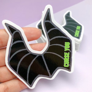 May include: A stack of stickers featuring a black and gray horn design with the text "CURSE YOU" in green. The stickers have a white border and a glossy finish. The design is inspired by a fantasy character.