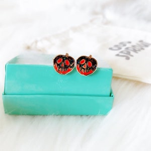 May include: A pair of gold-toned earrings featuring a red and black poison apple design. The earrings are in a small turquoise box.