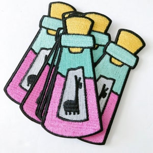 May include: A stack of five pink and turquoise embroidered iron-on patches featuring a cartoon llama in a bottle. The bottle has a yellow cork and a white label with the word "Llama" on it.