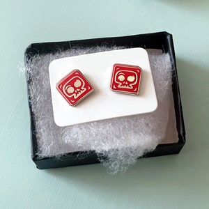 May include: A pair of red skull earrings with white details. The earrings are displayed on a white card with a black background.