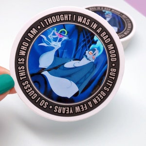May include: A round sticker with a black border and white background. The sticker features a cartoon image of Hades from Disney's Hercules, holding a martini glass. The text on the sticker reads "BUT IT'S BEEN A FEW YEARS. SO I GUESS THIS IS WHO I AM. I THOUGHT I WAS IN A BAD MOOD."