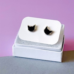 May include: A pair of silver cat stud earrings with black enamel. The earrings are displayed in a white box on a pink and gray surface.