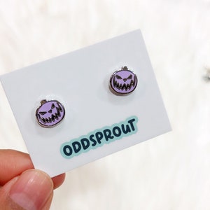 May include: A pair of purple pumpkin earrings with a spooky face design. The earrings are on a white card with the text "Oddsprout" printed on it.