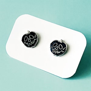 May include: A pair of black enamel earrings shaped like a poisoned apple with a skull face. The earrings are on a white background.