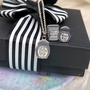 May include: A silver necklace with a charm of a glass jar labeled "Deadly Nightshade". The necklace comes in a black box with a white and black striped ribbon. The box also contains two other charms, one labeled "Frog's Breath" and the other labeled "Worm's Wort".