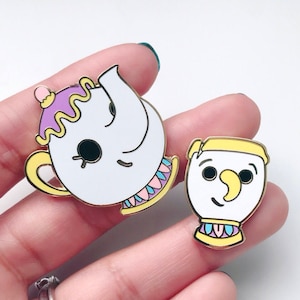 May include: Two enamel pins featuring characters from the Disney movie Beauty and the Beast. The first pin is a white teapot with a purple lid and gold trim. The second pin is a white cup with a yellow rim and gold trim.
