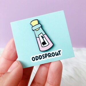 May include: A light blue and pink enamel pin of a bottle with a llama inside. The pin says "Oddsprout" on the bottom.
