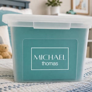 May include: A rectangular, semi-transparent plastic storage bin with a matching lid. The front of the bin features a white rectangular border containing the name MICHAEL in uppercase letters above the name thomas in lowercase letters.