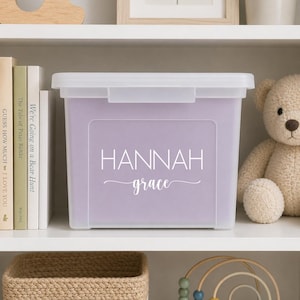 May include: A rectangular, light purple storage bin with a clear plastic lid. The front of the bin features the white text "HANNAH" in a clean, sans-serif font, with the name "grace" written below in a white, elegant cursive script.