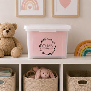 May include: A rectangular, semi-transparent white plastic storage bin with a lid, featuring a black circular leaf wreath graphic and the text "OLIVIA grace" printed on the front. The bin is positioned on a white shelf next to a brown teddy bear.