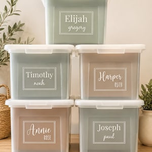 May include: Five rectangular plastic storage bins with white lids stacked in a pyramid. The bins are sage green and beige, each featuring a white rectangular label with names printed in a mix of serif and script fonts: Elijah Gregory, Timothy Noah, Harper Ruth, Annie Rose, and Joseph Paul.