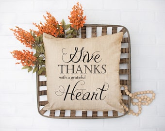 May include: A beige throw pillow with the text "Give Thanks with a grateful Heart" in black cursive lettering. The pillow is sitting in a wooden crate with a strand of wooden beads.