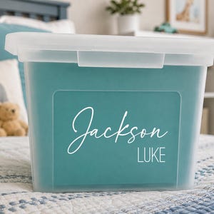 May include: A clear plastic storage bin with a lid, featuring a teal insert on the front. The insert displays the name Jackson in a white cursive script font, with the name LUKE printed below it in a clean, white, sans-serif capital font.