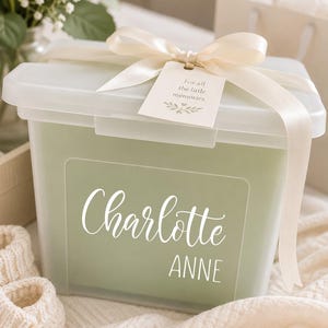 May include: A translucent plastic storage box with a sage green insert, personalized with the white text Charlotte ANNE. The lid is tied with a cream-colored satin ribbon and a small tag that reads For all the little memories.