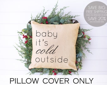 May include: A beige pillow cover with the text "baby it's cold outside" printed in black. The pillow cover is displayed on a white background with greenery and red berries.