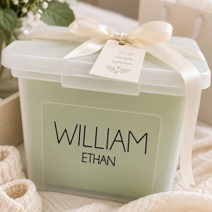 May include: A rectangular, translucent storage box with a sage green insert featuring the black text "WILLIAM ETHAN". The lid is secured with a cream-colored satin ribbon tied in a bow, holding a small tag that reads "For all the little memories".